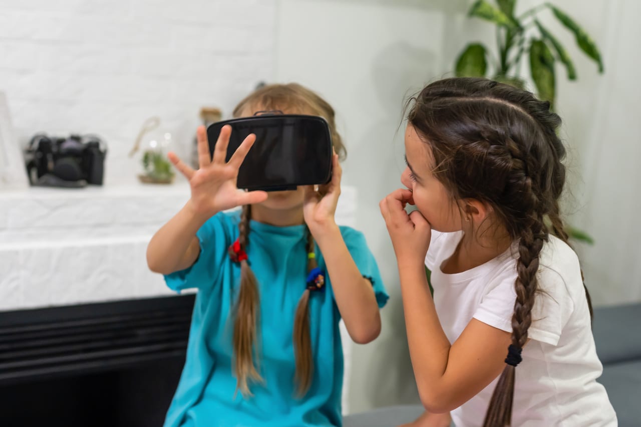 Two young girls, one holding a VR headset to her face while the other watches