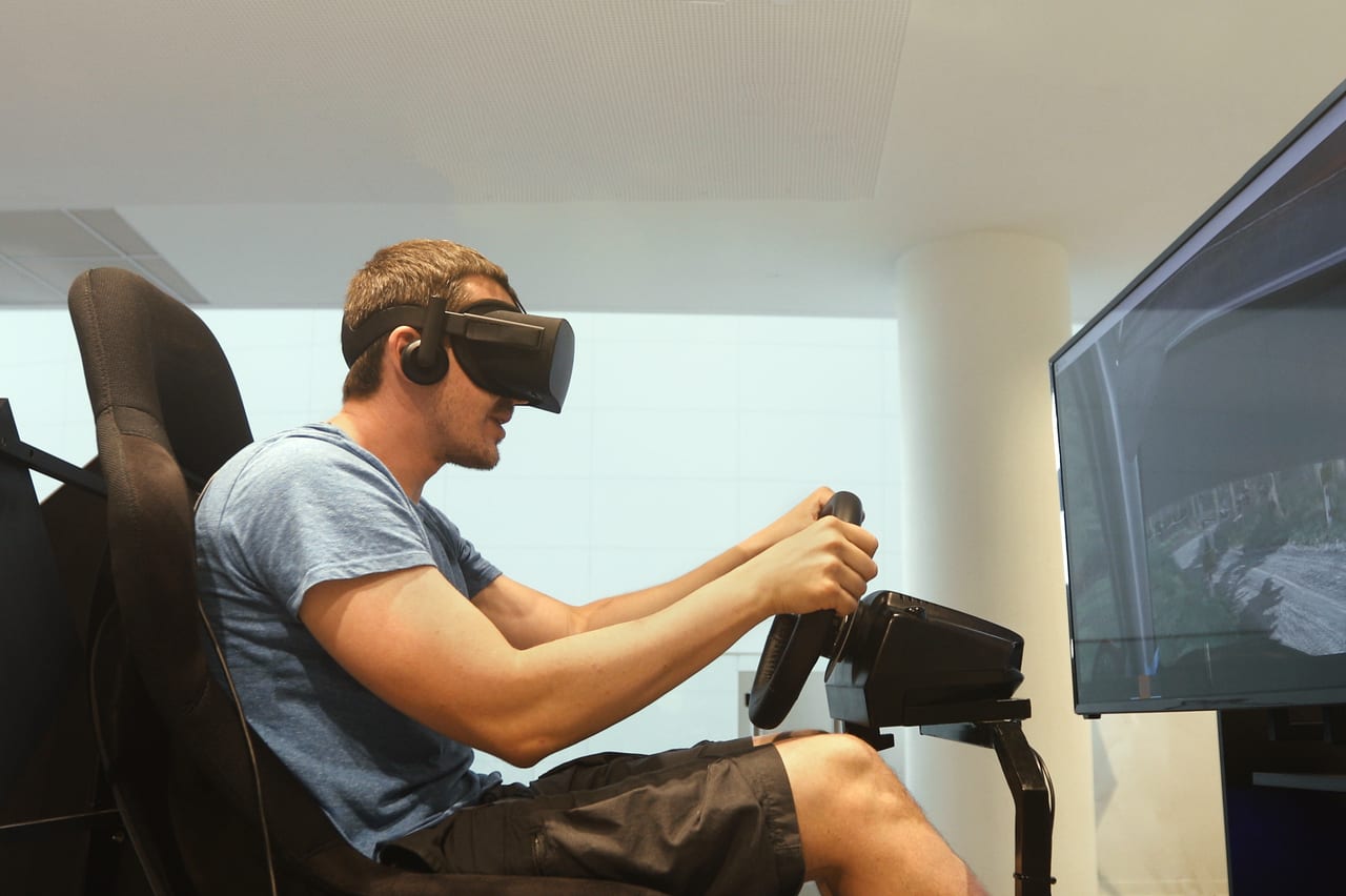 A man in a VR headset at a racing wheel setup, one hand on the shifter, a screen beside him
