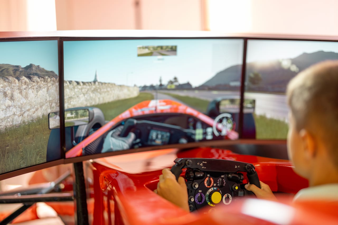 Over-the-shoulder view of a driver in a red racing simulator facing three track screens.