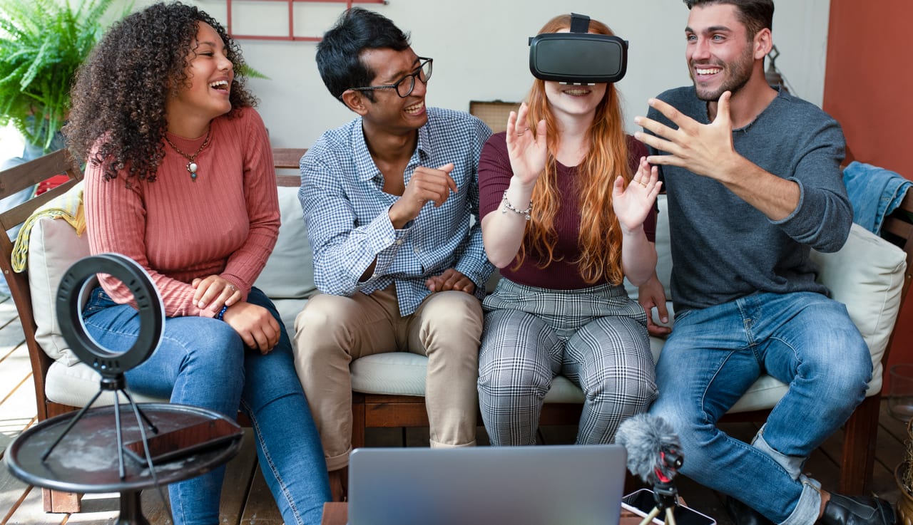 Four friends on a patio couch, one in a VR headset reaching out while the others laugh.