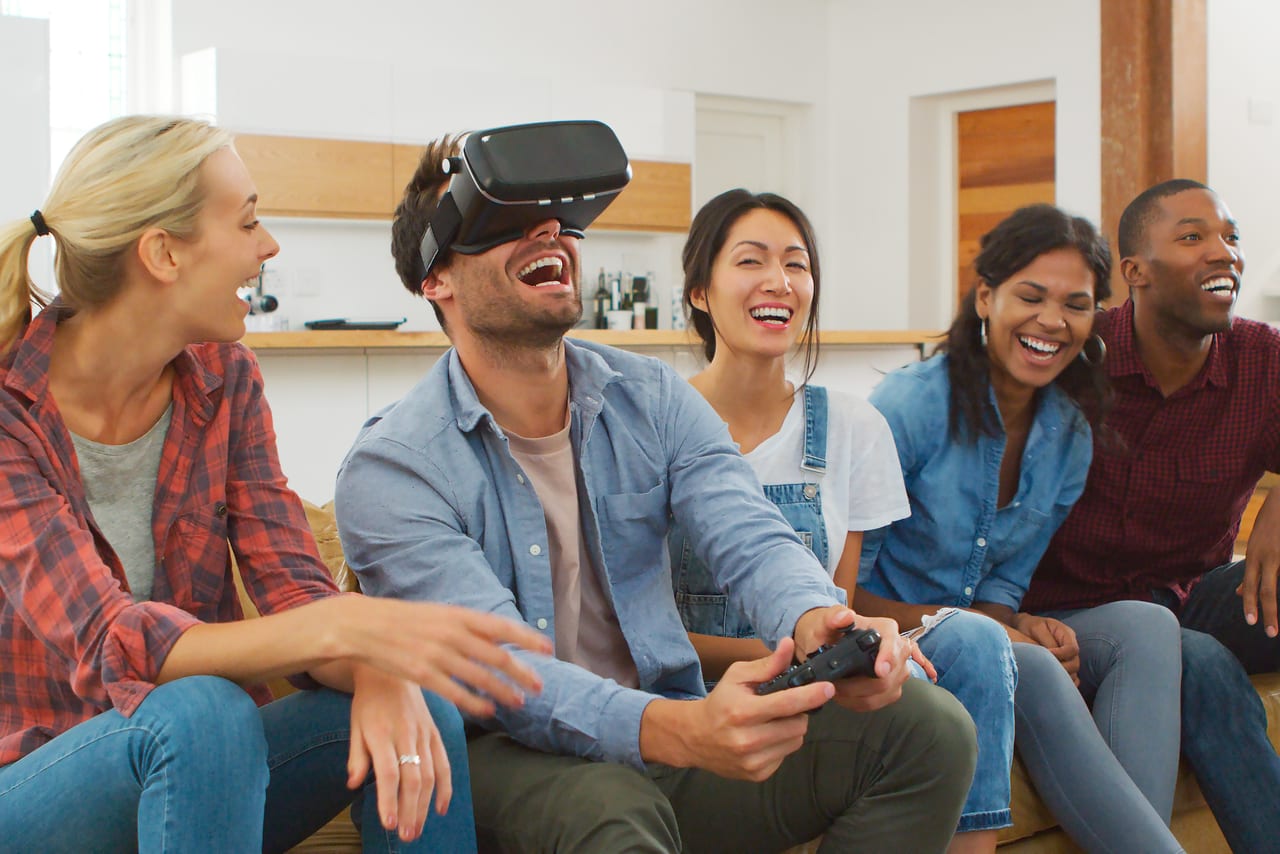 Five friends on a couch laughing while one of them plays in a VR headset