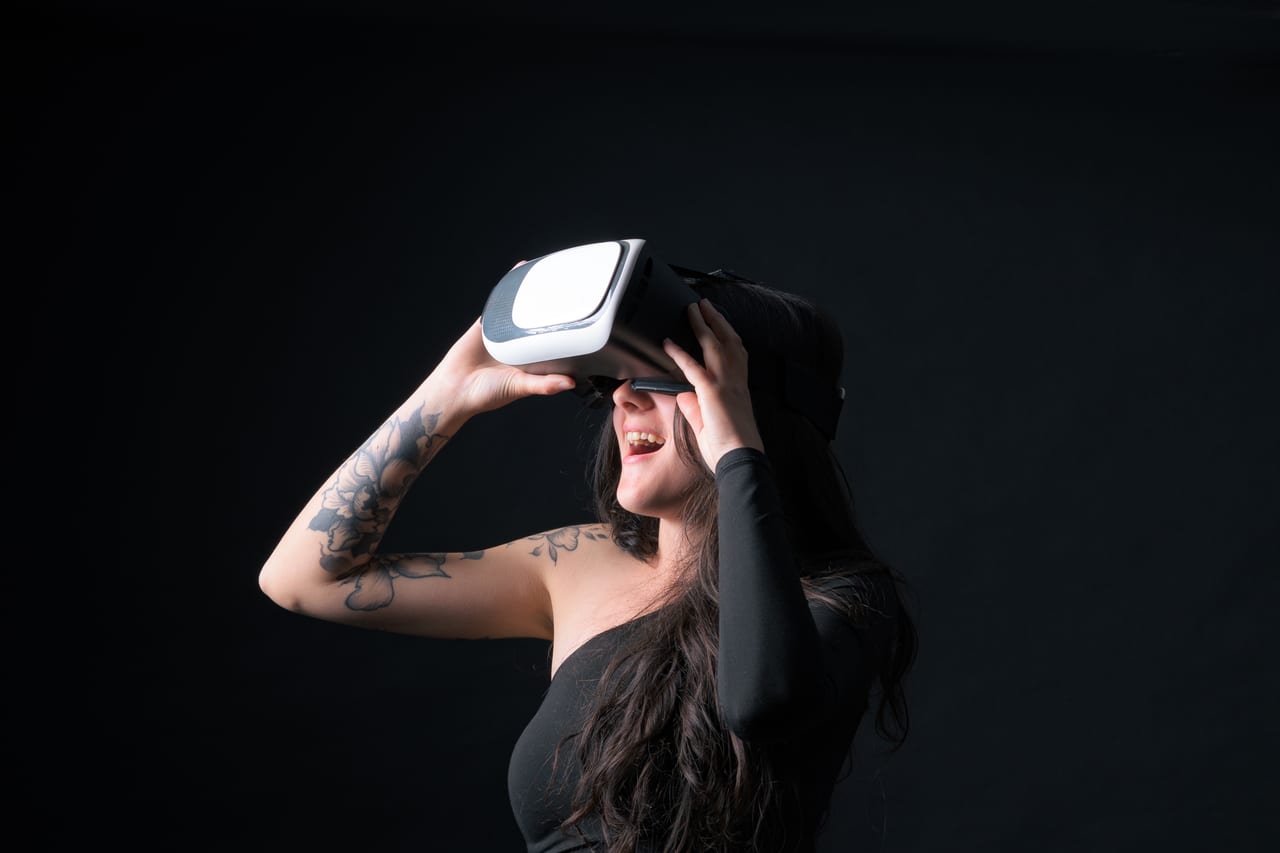 A woman holding a VR headset to her eyes, grinning, against a dark background.