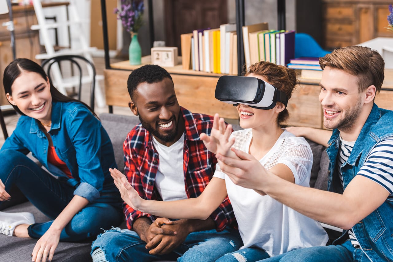Four friends on a couch, one in a VR headset reaching out while the others laugh.